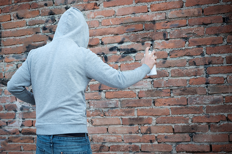 A person in a hooded sweatshirt is spray painting on a brick wall.