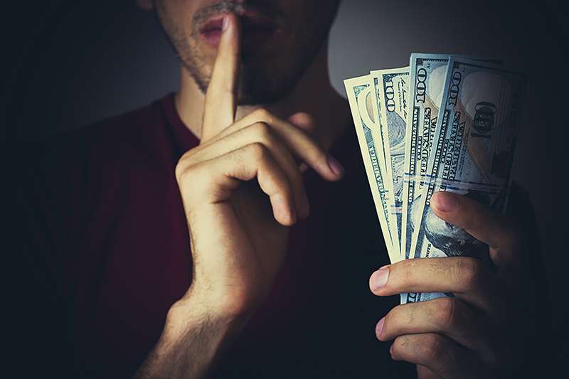 A person is holding a fan of cash with one hand while gesturing silence with a finger to their lips with the other.