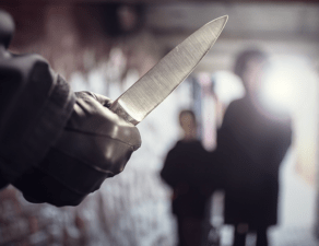 A person is ominously holding a knife with a pointed blade while another individual is blurred in the background, suggesting a threatening situation.