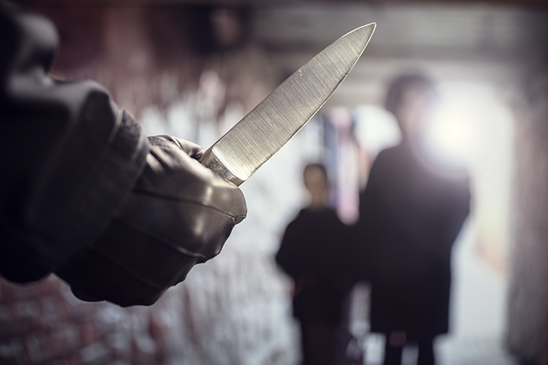 A person is ominously holding a knife with a pointed blade while another individual is blurred in the background, suggesting a threatening situation.