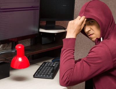 A person wearing a hooded sweatshirt is sitting at a desk with a computer, appearing stressed or concerned.