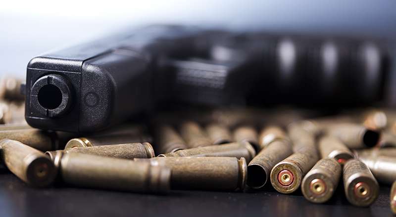 A handgun lies on a surface surrounded by scattered bullet casings, with a focus on the muzzle.