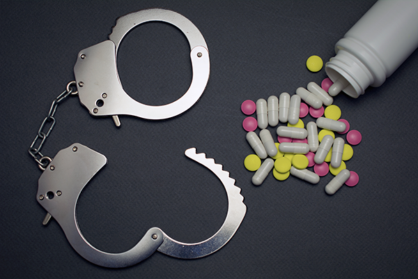 A pair of handcuffs next to spilled capsules and pills from an overturned bottle against a dark background.