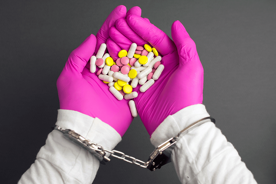 Hands in pink gloves handcuffed together, holding various pills against a dark background.