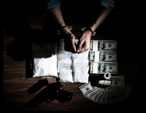 A person is surrounded by money, a gun, and what appears to be drugs, in a dimly lit setting, suggesting illegal activity.