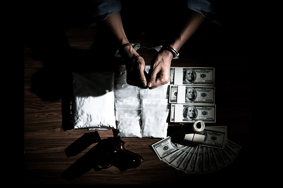 A person is surrounded by money, a gun, and what appears to be drugs, in a dimly lit setting, suggesting illegal activity.