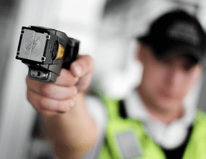 A security officer is aiming a handheld device, possibly for identification or scanning, towards the camera, with the focus on the device and the background blurred.