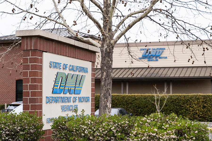 A sign for the Department of Motor Vehicles (DMV) in front of a building with trees and flowers in the foreground.