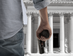 A person is covertly holding a handgun behind their back outside a building with classical columns.