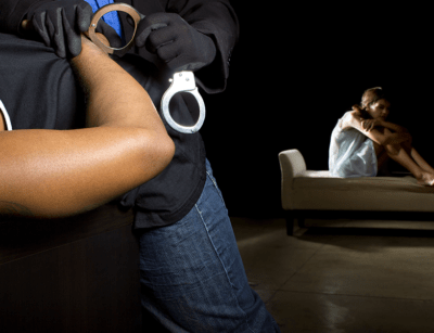 A person in a hoodie holding handcuffs appears to loom over a frightened woman seated on a couch in a dimly lit room.
