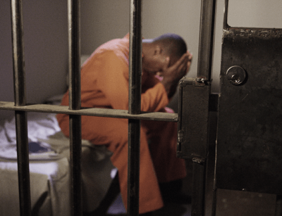 A person in an orange jumpsuit appears distressed behind the bars of a cell door.