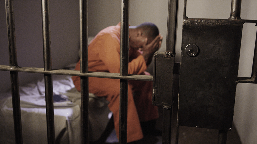 A person in an orange jumpsuit appears distressed behind the bars of a cell door.