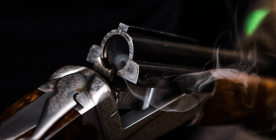 The image shows a close-up of a smoking double-barreled shotgun with intricate designs on the metal surfaces.