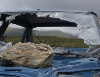 A large rock sits on the crushed roof and shattered windshield of a blue car with a grassy hill in the background.