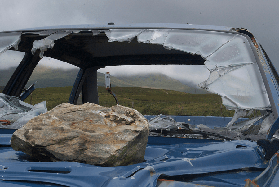 A large rock sits on the crushed roof and shattered windshield of a blue car with a grassy hill in the background.