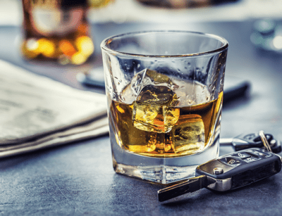 A glass of whiskey with ice next to a set of car keys and an unfolded newspaper on a dark surface.