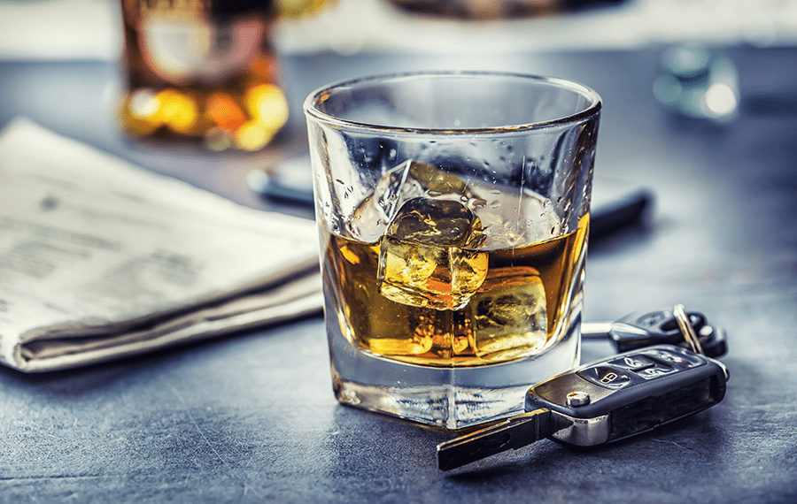 A glass of whiskey with ice next to a set of car keys and an unfolded newspaper on a dark surface.