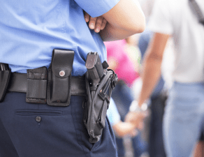 A close-up of a police officer's belt with equipment, including a holster with a gun, with people in the background.