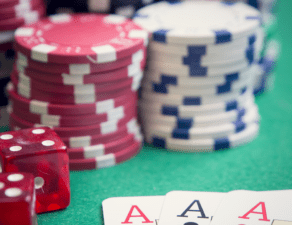 The image shows a set of four aces in the foreground with red dice and multiple stacks of poker chips in the background on a green surface.