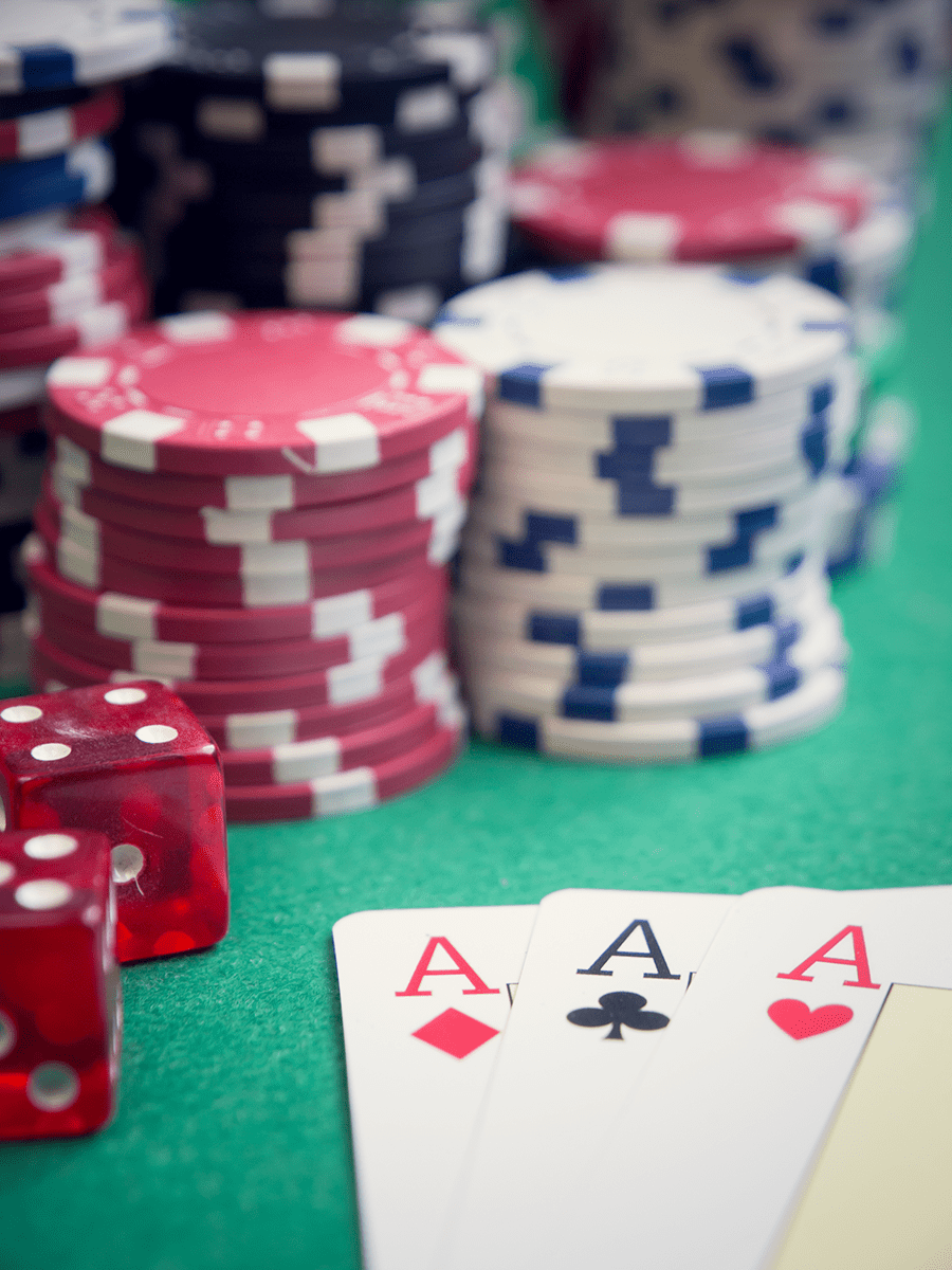 The image shows a set of four aces in the foreground with red dice and multiple stacks of poker chips in the background on a green surface.