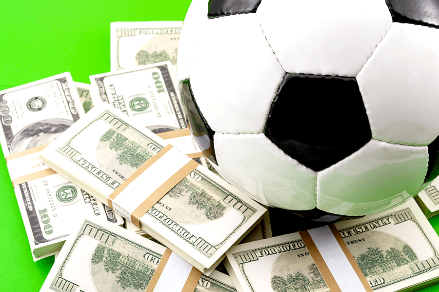 A soccer ball rests on a bed of US dollar bills against a green background, suggesting a concept of money in sports.