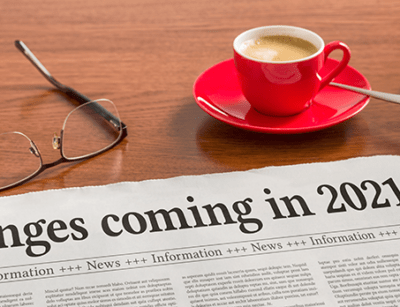 A newspaper headline reads Changes coming in 2021, alongside glasses, a red coffee cup, and a keyboard on a wooden desk.