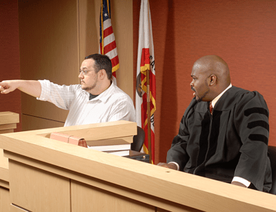 A person is testifying in a courtroom while pointing, and a judge listens attentively.