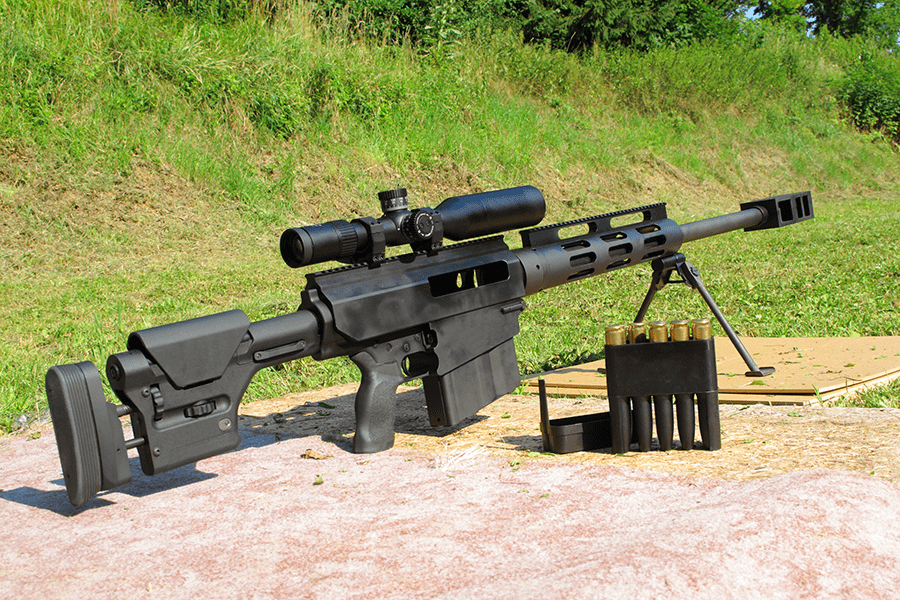A sniper rifle with a scope and ammunition laid out on a mat outdoors.