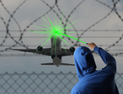A person in a blue hoodie is pointing a bright green laser at an airplane behind a barbed wire fence.