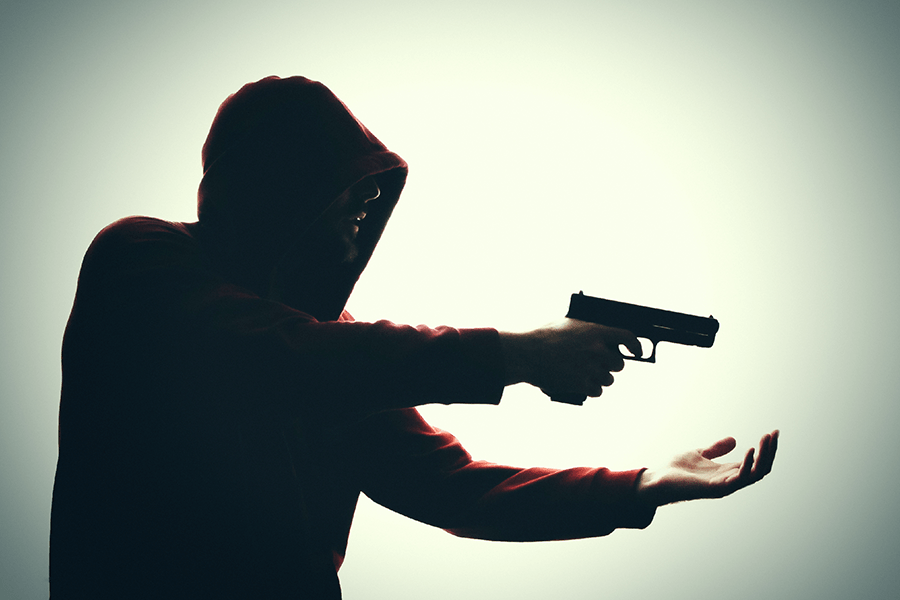 A silhouette of a person in a hooded top aiming a handgun to their right, against a backlit background.