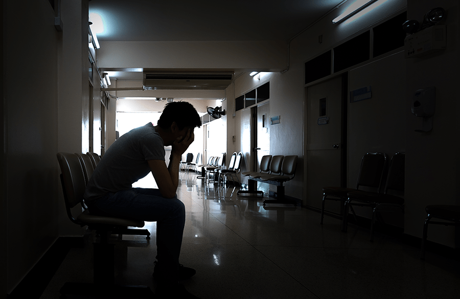 A person appears distressed, sitting with their head in their hands in a dimly lit, empty hospital corridor.
