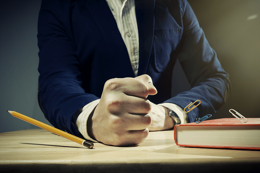 A person making a fist on a desk next to a closed notebook and a pencil.