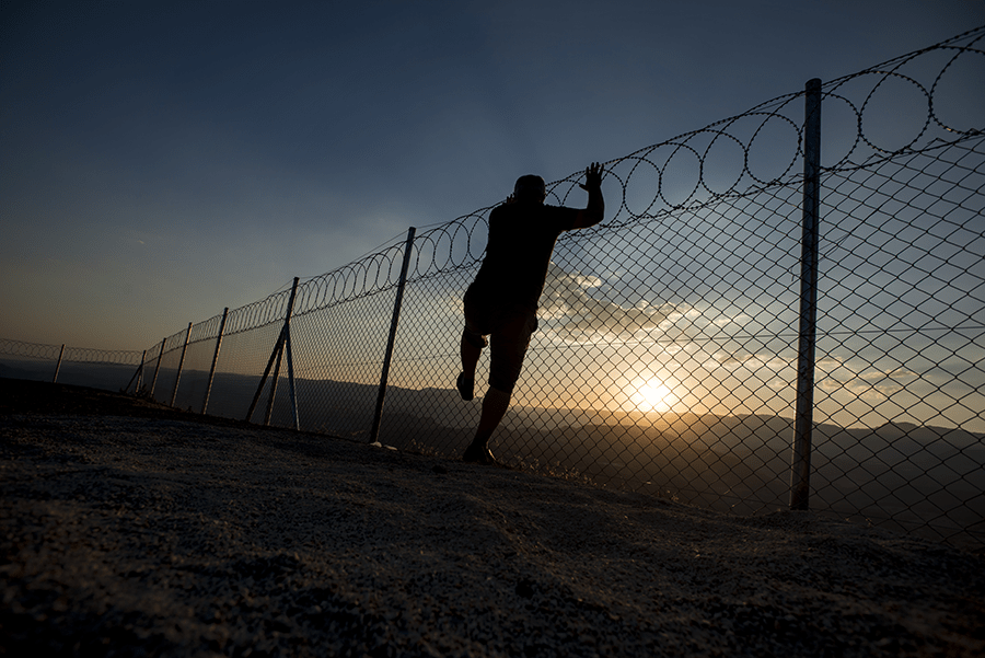 A person stands by a chain-link fence at sunset, with a silhouette effect against a vast, illuminating sky.
