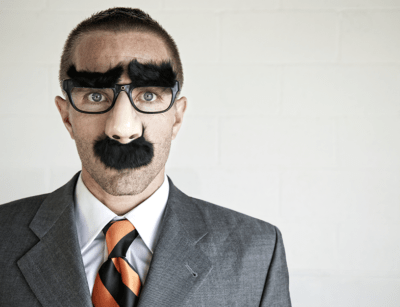 A man in a suit wearing a fake nose, mustache, and glasses disguise stands against a white brick wall background.