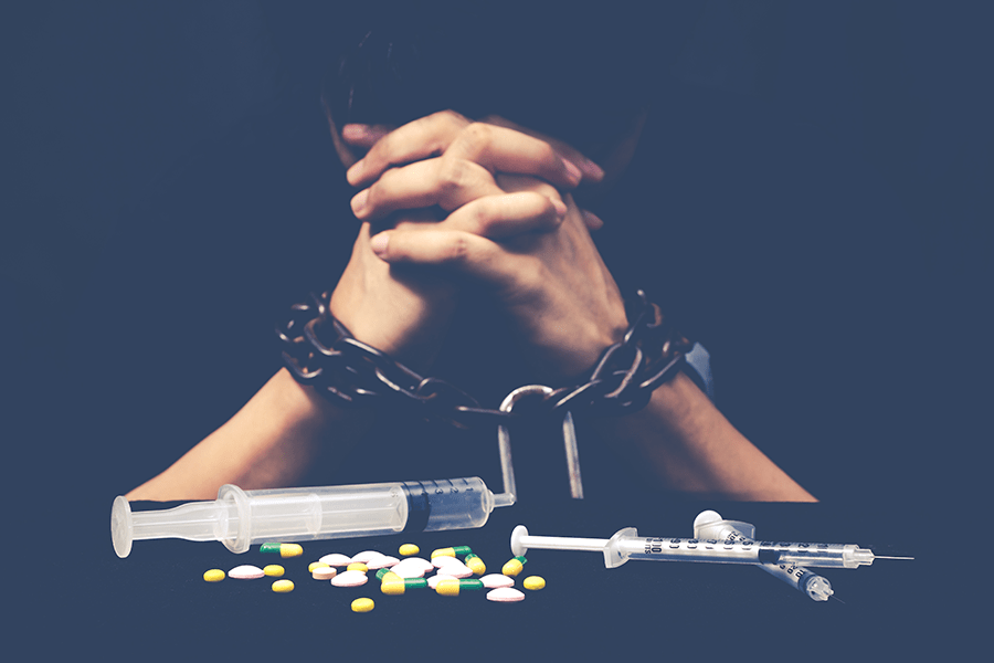 A person's chained hands represent addiction with scattered pills and syringes symbolizing substance abuse against a dark background.