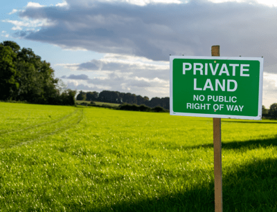 A sign reading PRIVATE LAND NO PUBLIC RIGHT OF WAY on a lush green field under a partly cloudy sky.