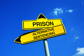 Two yellow directional signs against a blue sky, one reading PRISON and the other ALTERNATIVE SENTENCING.