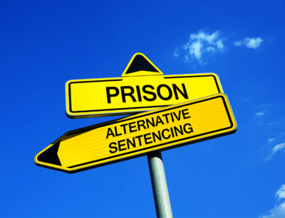 Two yellow directional signs against a blue sky, one reading PRISON and the other ALTERNATIVE SENTENCING.