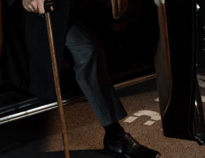 A person in formal attire is exiting a vehicle with a cane in hand.