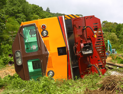 A derailed green and orange train lies tipped over beside the tracks in a lush green environment.