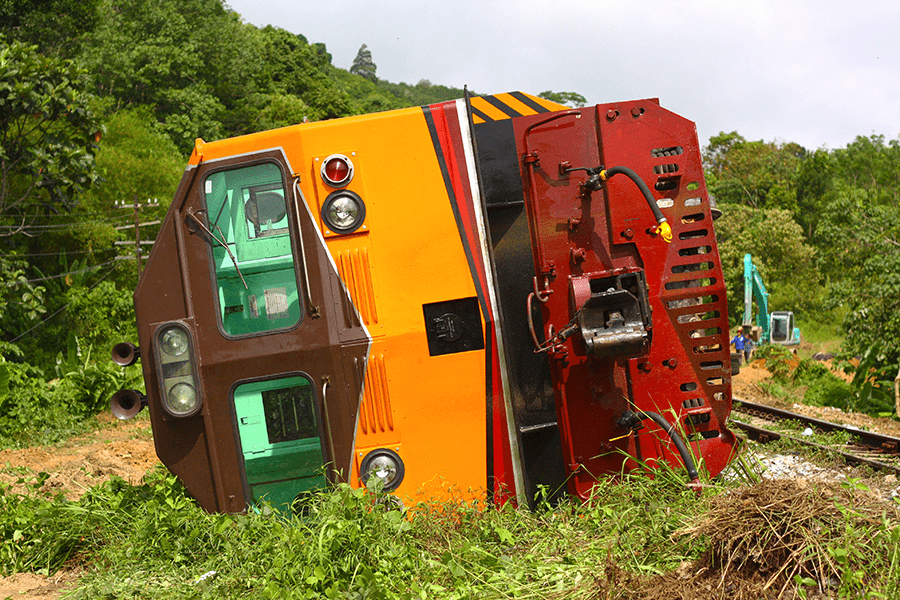 A derailed green and orange train lies tipped over beside the tracks in a lush green environment.