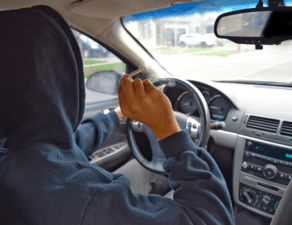 A person wearing a hood is driving a car while holding a cigarette.