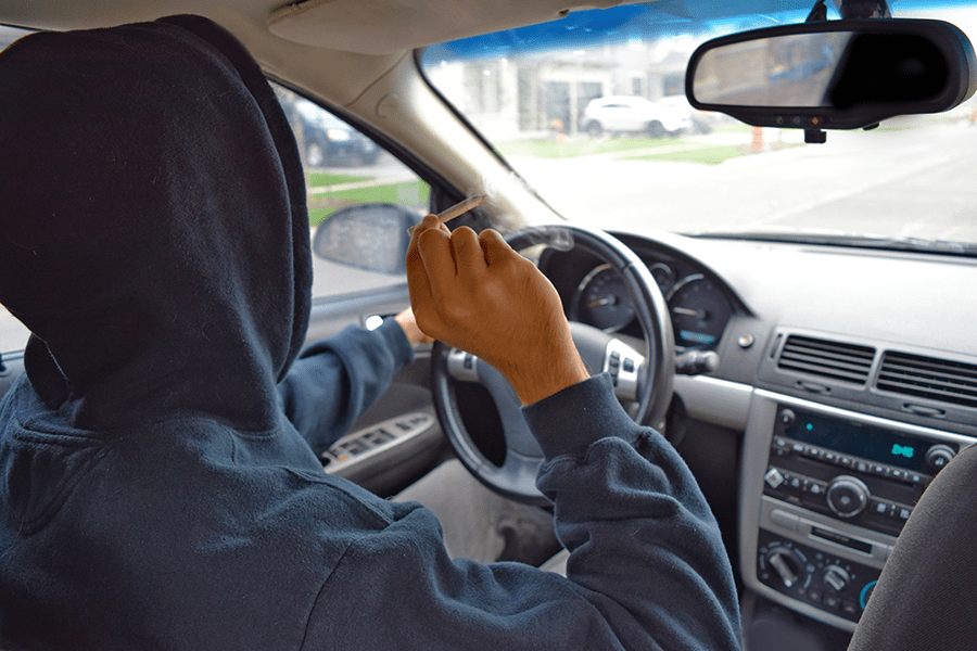 A person wearing a hood is driving a car while holding a cigarette.