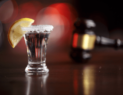A shot glass with a lemon slice and salt on the rim is in focus, with a blurred gavel in the background.