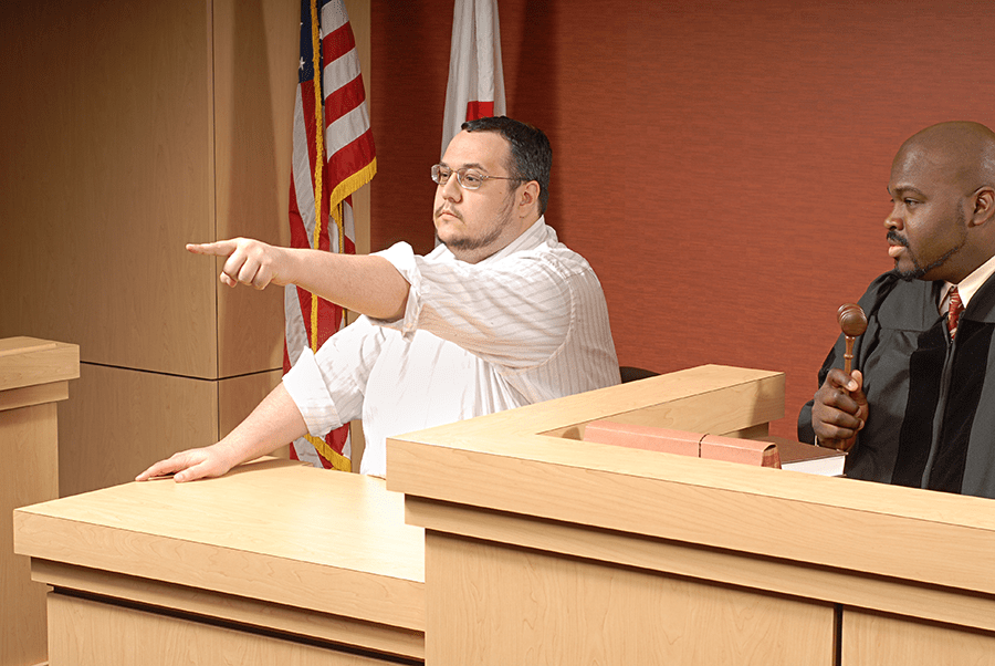 A man is testifying in court while pointing, with another man holding a gavel seated beside him.
