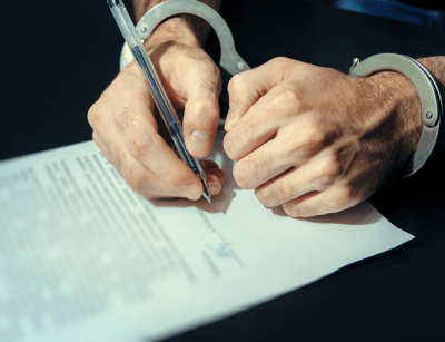 A person's handcuffed hands are signing a document on a dark surface.