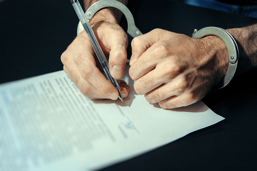 A person's handcuffed hands are signing a document on a dark surface.