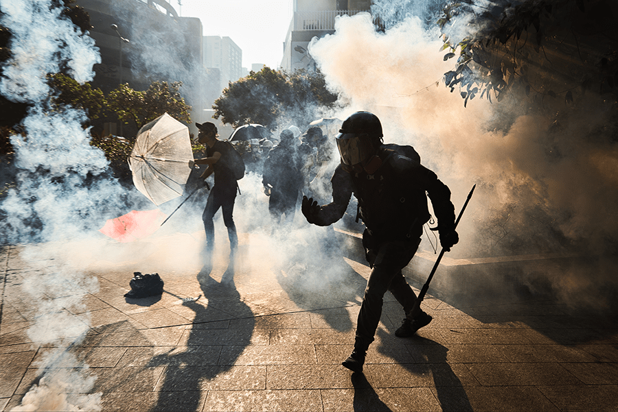 Protesters with umbrellas confront riot police amidst clouds of tear gas on an urban street.