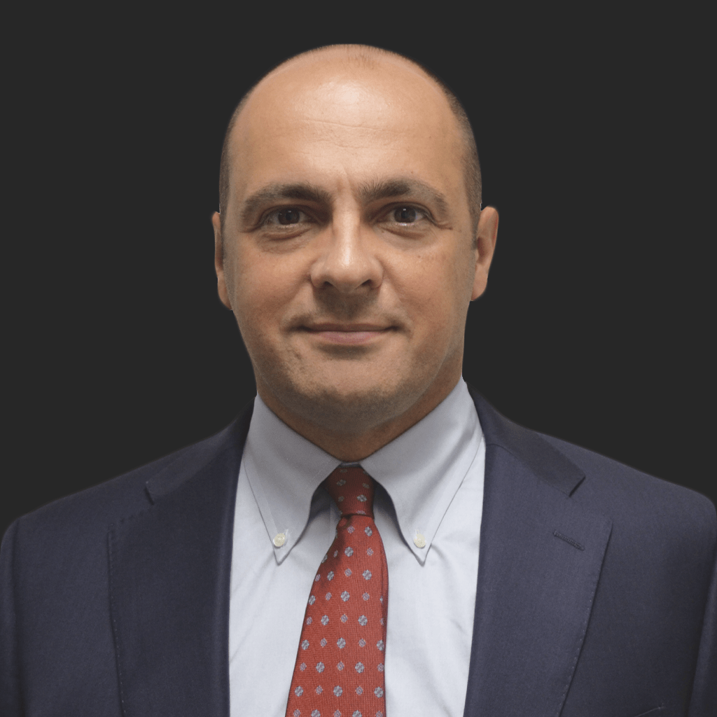 A man in a suit with a tie stands against a black background, displaying a neutral expression.