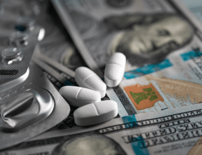 The image shows various pills and blister packs on top of US dollar bills, suggesting a theme of pharmaceutical expenses or healthcare costs.